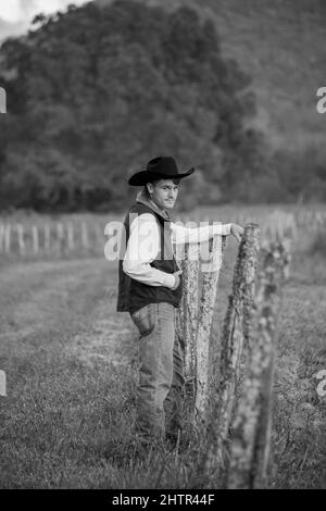 Photo verticale en niveaux de gris d'un homme portant une tenue de cow-boy et se tenant à côté d'une clôture dans la nature Banque D'Images