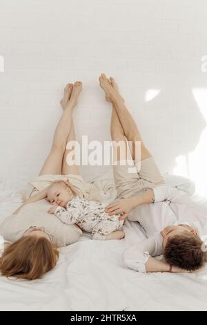 Jeune couple en vêtements blancs couchés avec des jambes soulevées droit sur le lit blanc, caressant mignon plenter bébé bleu-yeux. Banque D'Images