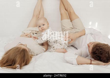 Jeune couple en vêtements blancs couchés avec des jambes soulevées droit sur le lit blanc avec bébé chubby bleu-eyed génial. Banque D'Images