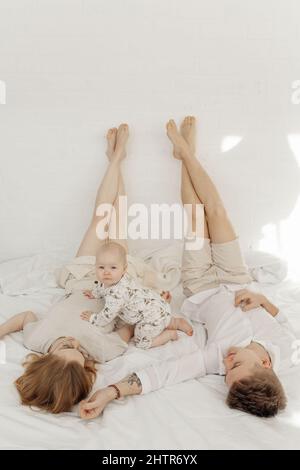 Jeune couple en vêtements blancs couchés avec les jambes soulevées droit sur le lit blanc avec le pump Blue-Eyed bébé. Banque D'Images