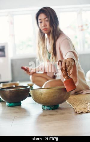 Femme jouant sur un bol de chant tibétain assis sur un tapis de paille à la maison Banque D'Images