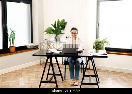 Jeune femme d'affaires concentrée dans les lunettes, assise au bureau, travaillant sur un ordinateur portable Banque D'Images