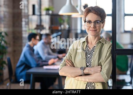 Portrait d'une femme d'affaires mûre dans des lunettes debout avec ses bras croisés et regardant la caméra avec ses collègues ayant une réunion en arrière-plan Banque D'Images