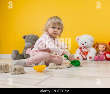 Une fille de deux ans en costume rose est assise sur le sol dans la salle de jeux et joue avec du sable cinétique beige Banque D'Images
