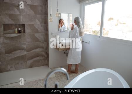 Belle femme asiatique avec de longs cheveux regardant dans le miroir dans la salle de bains Banque D'Images