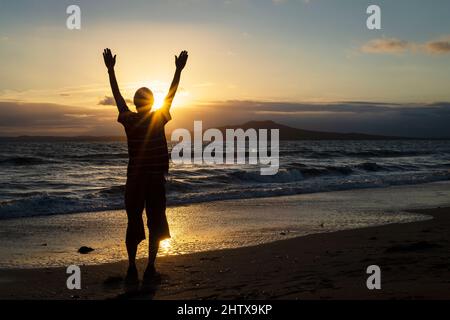 Silhouette d'un homme à bras ouverts saluant le soleil se levant sur l'île Rangitoto, Milford Beach, Auckland. Banque D'Images