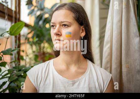 Une jeune fille ukrainienne triste avec le drapeau de l'Ukraine sur son visage regarde par la fenêtre. Le concept de la participation du peuple ukrainien au Banque D'Images