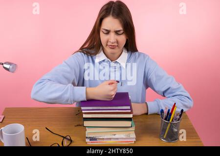 Belle blonde bouleversé jeune étudiant assis à la table en uniforme bleu d'université mettre son poing sur la pile de livres sur fond rose. Banque D'Images