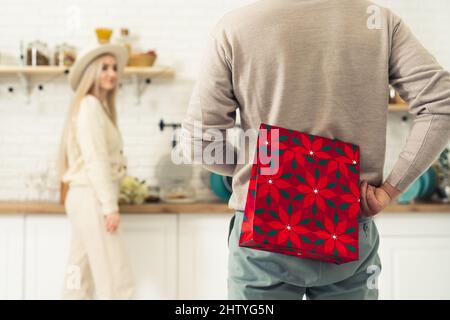 Homme européen debout et tenant un sac cadeau rouge derrière son dos pour son partenaire blond aux cheveux longs qui est debout dans la cuisine. Photo de haute qualité Banque D'Images