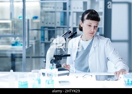Femme médecin en manteau blanc utilisant un comprimé numérique tout en travaillant avec des échantillons de virus pour créer un nouveau vaccin Banque D'Images