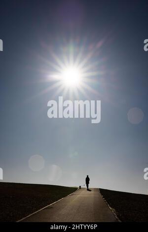 03 mars 2022, Schleswig-Holstein, Dagebüll: Une femme marche avec son chien sur la digue au bord du port sous un soleil éclatant. Photo: Christian Charisius/dpa Banque D'Images