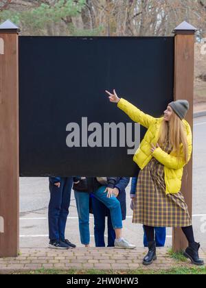 Une femme souriante vêtue de jeunes montre ses doigts sur un stand noir vide dans un parc municipal d'automne. Tableau vide pour écriture Banque D'Images