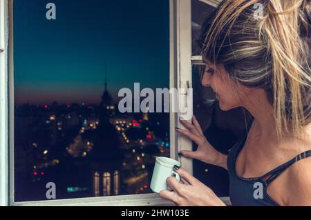 portrait de la jeune femme caucasienne souriante buvant un café regardant la ville de nuit par la fenêtre, concept de style de vie. Banque D'Images