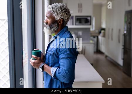 Homme barbu afro-américain tenant une tasse de café regardant par la fenêtre à la maison Banque D'Images