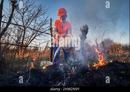 Écologiste pompier combattant le feu dans le champ avec le ciel du soir en arrière-plan. Homme écologiste tenant le seau et versant de l'eau sur l'herbe sèche brûlante. Concept de catastrophe naturelle. Banque D'Images