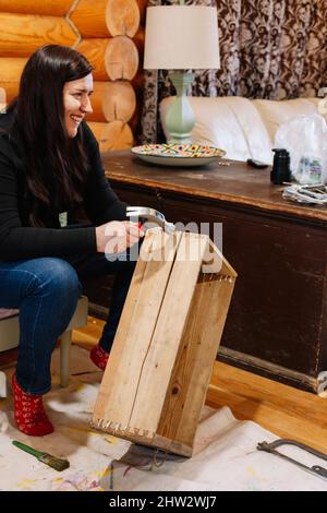 Femme d'âge moyen souriant fixant une ancienne boîte en bois avec un marteau dans l'atelier à la maison. Donner une vie nouvelle aux choses anciennes. Réutilisation des anciens. Actions durables pour Banque D'Images