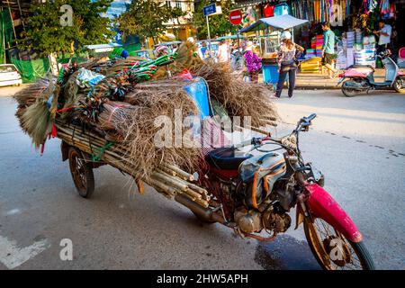 Une moto rouge avec un chariot accroché à l'arrière transportant du bois, du bambou, des balais, pour la livraison, est garée dans la rue en face de l'ancien marché. Banque D'Images