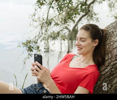 Femme souriante se penchée sur une branche d'arbre et regardant un smartphone Banque D'Images