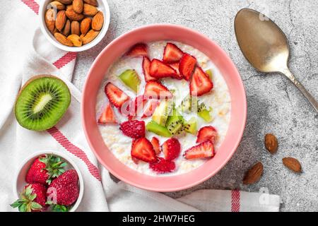 Porridge de flocons d'avoine avec tranches de kiwi, fraises, amandes dans un bol rose, cuillère, serviette avec rayures rouges sur fond de béton Vegan concept alimentaire Hea Banque D'Images