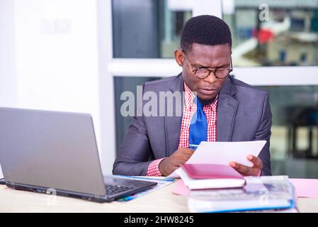 homme d'affaires adulte concentré assis devant un ordinateur portable et lisant le rapport sur papier au bureau. Banque D'Images