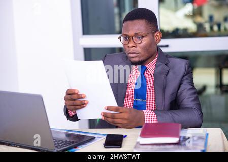 Un homme d'affaires sérieux adulte assis devant un ordinateur et lisant un rapport papier au bureau. Banque D'Images