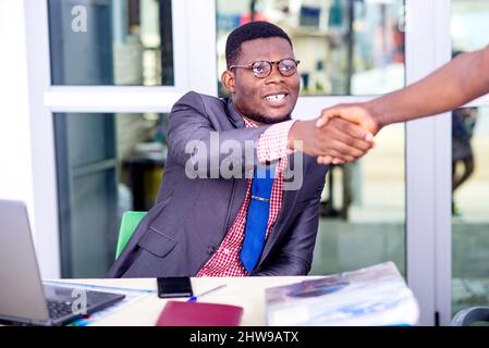 jeune homme d'affaires beau en veste assis au bureau en serrant la main avec son partenaire souriant. Banque D'Images