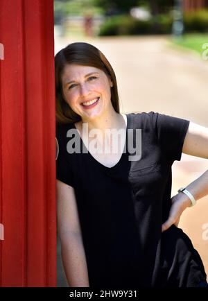 Une jeune femme se penche contre une cabine téléphonique rouge dans le centre-ville de Collierville, Tennessee. Elle a sur un tee-shirt noir et est souriante. Banque D'Images