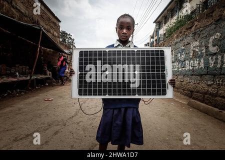 Une jeune fille d'école a vu porter un panneau solaire par les rues de Kibera. Dans les bidonvilles de Kibera, la plupart des résidents, en particulier les vendeurs de rue, les écoles privées, les organisations non gouvernementales, les éclairages de rue et les écoles, utilisent tous le système d'énergie solaire bon marché qui est une source d'énergie plus fiable et bon marché. Cela a facilité le recyclage de la plupart de l'énergie électrique consommée par les résidents et a également contribué à réduire le nombre élevé de décès dus aux chocs électriques et aux incendies de taudis quotidiens. Banque D'Images