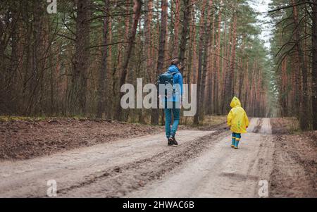 Maman et enfant marchant le long de la route de la forêt après la pluie dans les manteaux de pluie ensemble, vue arrière Banque D'Images