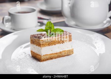 Morceau rectangulaire de Tiramisu avec une feuille de menthe sur une plaque blanche. Dans une clé faible, dans l'arrière-plan une tasse de thé et une bouilloire, vaisselle. Au service, la serv Banque D'Images