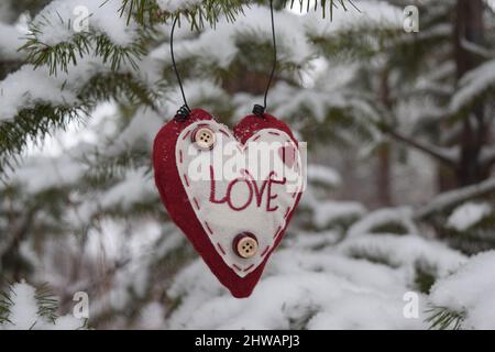 Décoration en forme de coeur faite de tissu et de boutons avec le mot amour accroché sur le pin recouvert de neige. Banque D'Images