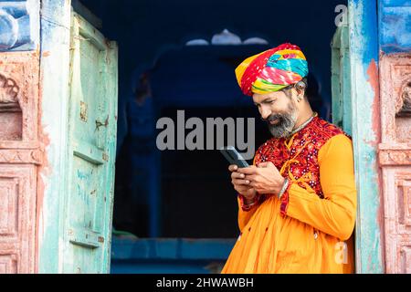 Un homme adulte originaire du nord de l'Inde, vêtu d'une tenue colorée et d'un turban avec un smartphone, souriant, appelant un homme traditionnel sur un smartphone android. Banque D'Images