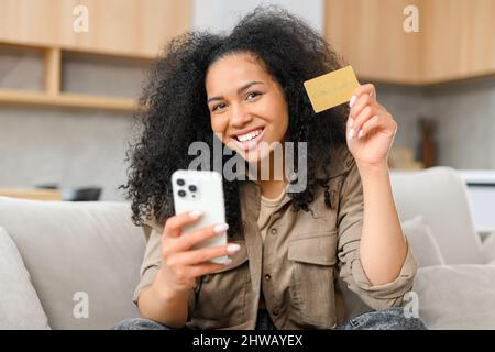 Une jeune femme joyeuse utilisant l'application d'achat sur smartphone pour acheter en ligne, en tenant sa carte de crédit et en entrant ses données bancaires, s'assoit sur un canapé dans une atmosphère détendue dans le salon de la maison Banque D'Images