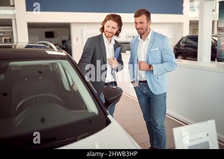 Un vendeur présente un nouveau modèle à un client dans une atmosphère agréable dans un salon de l'auto. Voiture, boutique, achat Banque D'Images