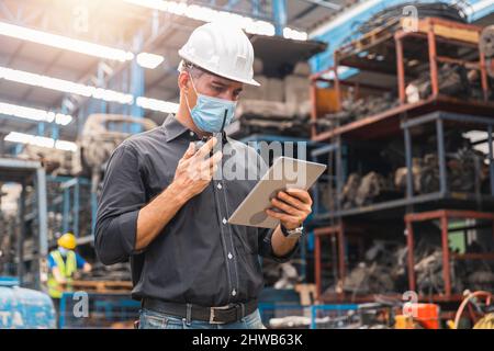 Inspecteur contremaître portant un masque facial ou chef d'ingénieur regardant la tablette et le contrôle travaillant par radio en usine Banque D'Images