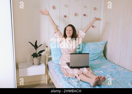 Jeune femme d'affaires heureuse se trouve dans un lit en pyjama dans une maison confortable. Travailler en ligne, étudier à domicile avec un ordinateur portable. Indépendant. Rituels du matin. Banque D'Images