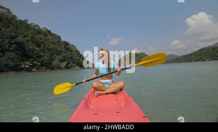 Belle femme blonde sportive avec des lunettes de soleil rangées de plastique rose canoë le long de l'eau de mer contre les collines vertes et le ciel bleu. Voyager dans les pays tropicaux. Une jeune fille sportive navigue en kayak dans l'océan. Banque D'Images