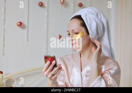 Jeune femme heureuse avec une serviette sur la tête faisant des procédures de soin de peau de visage. Lit en pyjama dans une maison confortable avec téléphone. Technologie. Médias sociaux Banque D'Images