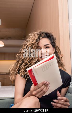 jeune femme caucasienne latine vénézuélienne avec des curlers souriant avec un livre dans la main regardant la caméra, assis sur le canapé à la maison. Banque D'Images