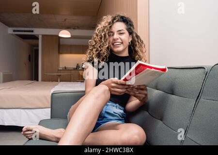 jeune fille caucasienne assise sur le canapé dans son appartement avec la cuisine et son lit derrière elle, tenant un livre souriant souriant souriant souriant regardant le chameau Banque D'Images