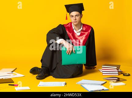Portrait du jeune homme, étudiant en chapeau de graduation et robe avec dossier assis sur le sol isolé sur fond jaune de studio Banque D'Images
