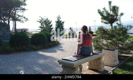 Jeune fille enoyant des buissons verts d'été et la mer.Vue arrière d'une femme avec beaucoup de tresses assis sur un banc en marbre blanc en face d'un magnifique emba Banque D'Images