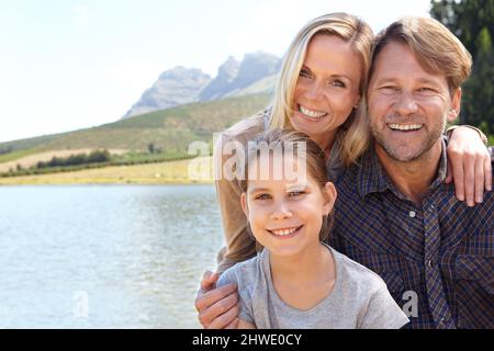 Des souvenirs qui durent toute une vie. Portrait d'une famille heureuse de trois personnes assises ensemble au bord d'un lac. Banque D'Images