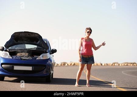 Les shes ont besoin d'une assistance routière. Une belle jeune femme debout avec sa voiture en panne en train de dételer un tour sur le côté de la route. Banque D'Images