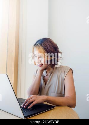 Femme asiatique, freelance avec cheveux courts tendance et regarder l'écran tout en travaillant avec un ordinateur portable sur un bureau rond en bois sur un mur blanc Banque D'Images