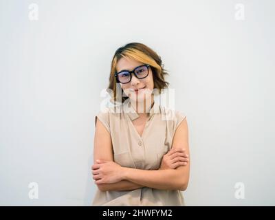 Souriante femme asiatique cheveux courts, style tendance en lunettes noires et chemise sans manches beige posant avec des bras croisés, regardant l'appareil photo sur blanc Banque D'Images