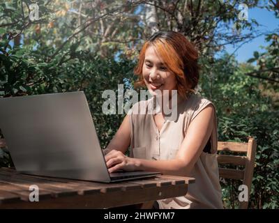 Beau portrait de femme asiatique heureuse avec cheveux courts tendance, regardant un écran d'ordinateur portable argenté sur un bureau en bois avec un sourire relaxant Banque D'Images