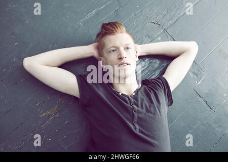 Perdu dans un rêve de jour. Un homme aux cheveux rouges allongé sur le sol et regardant vers le haut avec un regard réfléchi sur son visage. Banque D'Images