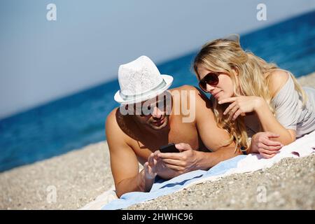 Garder le contact n'a jamais été aussi simple. Photo d'un jeune couple utilisant un téléphone portable tout en bronzant sur une plage. Banque D'Images