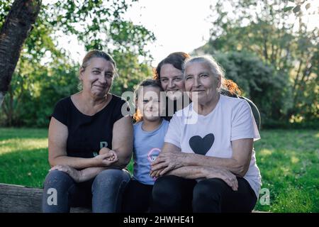 Quatre générations de femmes d'une famille assis sur le bois dehors regardant la caméra sourire dans la journée avec de la verdure en arrière-plan. Passer du temps ensemble Banque D'Images